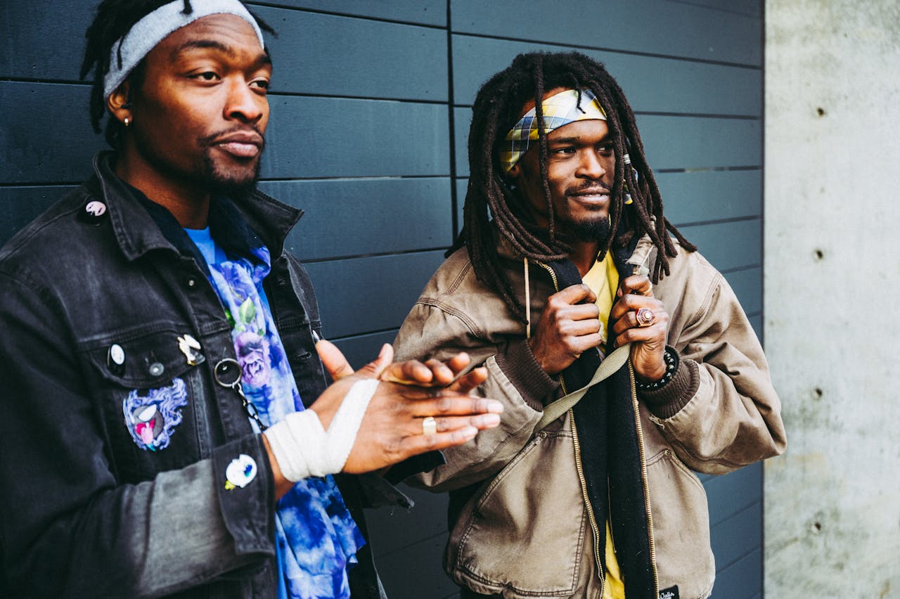Two African American men in stylish casual streetwear stand confidently against a modern wall.